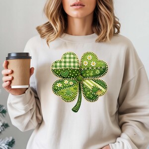 May include: A cream-coloured sweatshirt with a large, patchwork-style four-leaf clover design. The clover features various green patterns, including gingham, polka dots, and stripes, with small white daisy accents. A heart is stitched on one leaf.