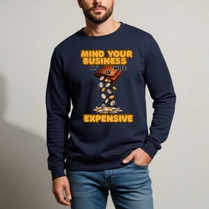 May include: Navy blue sweatshirt with the text "MIND YOUR BUSINESS MY WIFE IS EXPENSIVE" in yellow and brown. The graphic shows a brown wallet with coins pouring out, suggesting high expenses. The sweatshirt is a casual wear item.