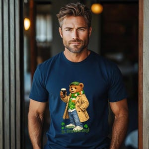 Puede incluir: Camiseta azul marino con un oso de dibujos animados con gorro verde, bufanda y abrigo beige, sosteniendo una pinta de cerveza oscura. La camiseta tiene el texto "Happy St. Patrick's Day!" con tréboles.