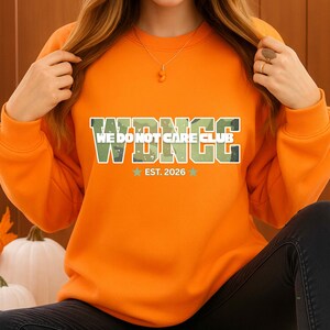 以下が含まれることがあります： オレンジ色のスウェットシャツ。迷彩柄で「WDNCC WE DO NOT CARE CLUB」の文字がプリントされています。テキストの下には、2つの星が付いた「EST. 2026」があります。スウェットシャツは無地のオレンジ色です。