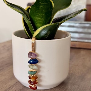 May include: A gold-toned metal clip ornament featuring a vertical row of seven polished, multi-colored gemstone chips in shades of purple, blue, light green, green, yellow, orange, and red, attached to the rim of a plain, matte white ceramic pot.