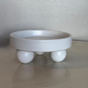 May include: A speckled white ceramic dish with a rounded rim, supported by three spherical feet. The dish has a shallow bowl shape and a speckled texture, creating a minimalist aesthetic. The neutral color and simple design make it versatile for various uses.