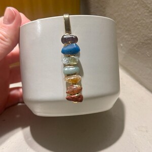 May include: A white ceramic mug with a gold-colored metal accent. A vertical row of colorful gemstones is attached to the mug. The gemstones include purple, blue, green, yellow, and brown stones. The mug is held by a hand.