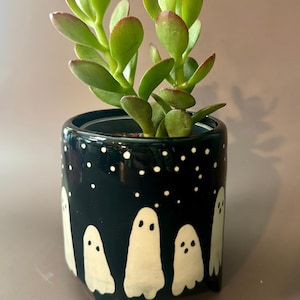 May include: A small, black ceramic plant pot featuring a ghost design. The pot contains a green succulent plant. The ghosts are white with simple shapes, and the background is dotted with white stars. The pot is approximately 10 cm tall.