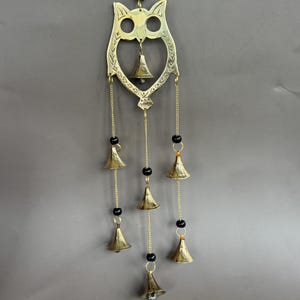 May include: A brass owl-shaped wind chime with seven small bells. The owl has cut-out eyes and a heart-shaped body. The bells hang from gold chains and are accented with black beads. The wind chime is designed for indoor or outdoor use.