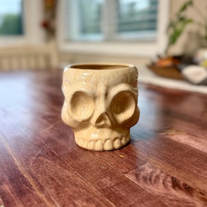 May include: A ceramic skull-shaped planter or mug with a glossy, cream-colored glaze featuring subtle tan and beige marbling throughout. The object has deep, hollowed-out eye sockets, a defined nasal cavity, and a row of teeth along the bottom edge.