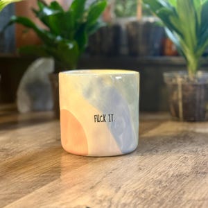 May include: A cylindrical ceramic mug featuring a hand-painted abstract design with soft pastel washes of peach, light blue, and sage green. The text FUCK IT. is printed in a simple, black, sans-serif font on the front center of the mug.