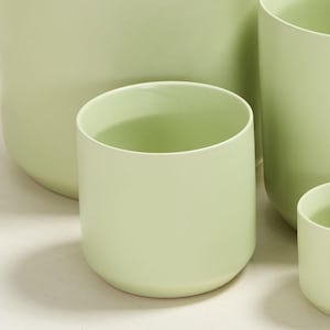 Puede incluir: Colección de macetas de cerámica verde claro de varios tamaños. Las macetas tienen un acabado liso y mate y una forma cilíndrica simple. La maceta más grande tiene aproximadamente 30 cm de diámetro.