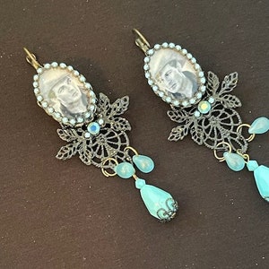 Op de afbeelding: Een paar oorbellen in antieke stijl. Elke oorbel heeft een ovaal portret, omringd door kleine blauwe kralen. Daaronder een filigraan ontwerp met een druppelvormige turquoise kraal. Bronzen afwerking.