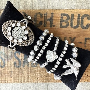 May include: A collection of silver-toned bracelets with sparkling embellishments and charms, including a bracelet with a bird pendant and another with the text "CONTINUE TO GROW". The bracelets are displayed on a black velvet cushion.