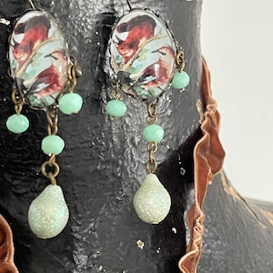 Puede incluir: Un par de pendientes colgantes con un diseño de inspiración vintage. Cada pendiente presenta un dije redondo de vidrio con una imagen colorida de pájaros y una ardilla. Los dijes están unidos a una cadena con cuentas de vidrio verde y una cuenta de lágrima nacarada.