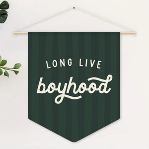 May include: A dark green banner with vertical stripes and the words "LONG LIVE boyhood" in white script. The banner is suspended from a wooden rod and white string. Green leafy branches are visible on the left side.