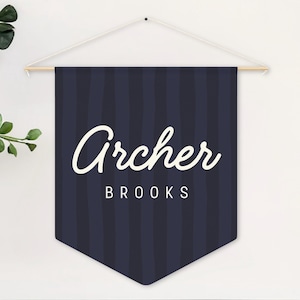 May include: A navy blue pennant banner with vertical stripes and the name "Archer" in white script. Below the name is the word "Brooks" in a smaller, block font. The banner is suspended from a wooden dowel.