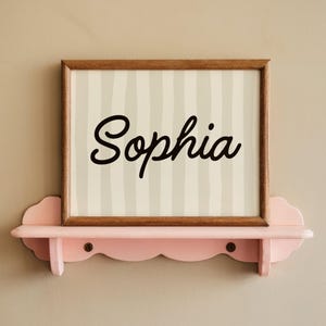 May include: A rectangular wooden frame containing a print with vertical light gray stripes and the name Sophia written in black script, resting on a scalloped light pink wall shelf with two visible dark circular screw heads.