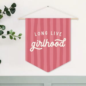 May include: A pink pennant banner with vertical stripes and the words "LONG LIVE girlhood" in white script. The banner is suspended from a wooden dowel and white string. Green leafy branches are visible on the left.