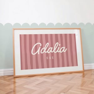 May include: A rectangular framed print featuring a dusty rose background with vertical stripes in a slightly darker shade. The name Adalia is written in a large white cursive script, with the name SUE printed below in a smaller white sans-serif font.
