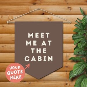 May include: A brown banner with the text "MEET ME AT THE CABIN" in white letters. A small, round, peach-colored area with the text "YOUR QUOTE HERE" is in the lower left corner. The banner hangs on a wooden background.