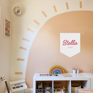 Pode incluir: Um quarto de brincar infantil com uma faixa branca que diz "Stella June" em escrita rosa. A parede &eacute; pintada num esquema de cores p&ecirc;ssego e branco com um design de arco-&iacute;ris. Uma mesa branca cont&eacute;m brinquedos, incluindo um arco-&iacute;ris de madeira e um piano de brinquedo.
