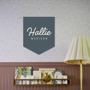 Pode incluir: Uma faixa de bandeira azul-escura com o nome "Hallie Madison" em letras brancas e em bloco. A faixa est&aacute; pendurada em um bast&atilde;o de madeira e barbante. Uma estante com livros infantis e uma l&acirc;mpada com uma sombra creme est&atilde;o no fundo.