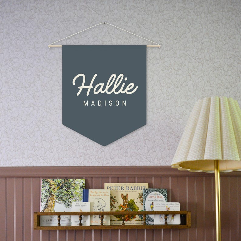 Pode incluir: Uma faixa de bandeira azul-escura com o nome "Hallie Madison" em letras brancas e em bloco. A faixa est&aacute; pendurada em um bast&atilde;o de madeira e barbante. Uma estante com livros infantis e uma l&acirc;mpada com uma sombra creme est&atilde;o no fundo.