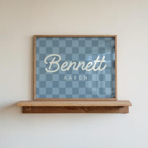 May include: A framed print with the name "Bennett" in a cursive font and the name "Aaron" in a smaller font, set against a blue and white checkered background. The frame is a light brown color, and the print is displayed on a wooden shelf.