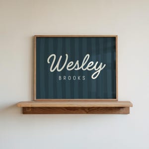 May include: A framed sign with the name "Wesley Brooks" in white script lettering on a dark blue striped background. The sign is displayed on a wooden shelf against a white wall. The frame is made of light brown wood.