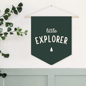 May include: A dark green banner with the words "little EXPLORER" in white, and a small white tree graphic. The banner is suspended from a wooden rod and white string. Green leafy branches are visible on the left side.