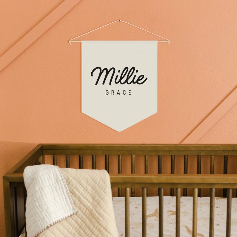 Pode incluir: Uma faixa creme com o nome "Millie" e a palavra "Grace" em escrita preta. A faixa est&aacute; pendurada em uma haste e corda douradas contra uma parede cor de p&ecirc;ssego. Uma cama de madeira &eacute; vis&iacute;vel abaixo.