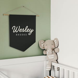 Pode incluir: Uma faixa preta com o nome "Wesley Brooks" em escrita branca est&aacute; pendurada em uma parede verde. Um elefante de pel&uacute;cia com orelhas listradas e um bolso est&aacute; ao lado de um ber&ccedil;o branco.
