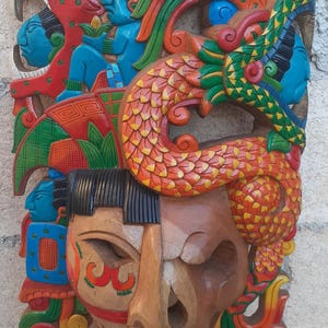 May include: A hand-carved wooden wall mask featuring a central face with half natural wood and half painted details. The upper section displays a vibrant, multi-colored relief of a serpent and human figures in red, blue, green, and yellow paint.
