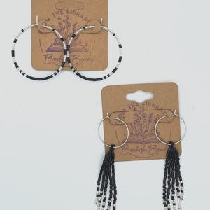 May include: Two pairs of hoop earrings. One pair has black and silver beads around the hoop. The other pair has black beaded fringe hanging from the bottom. Both pairs are displayed on brown cards with the text "From the Library Bookish Beads".