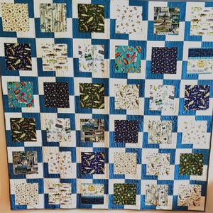May include: A handmade quilt with a fishing theme. The quilt features a patchwork design with squares of various fabrics, including fish, fishing rods, and lake scenes. The squares are set against a blue and white checkered background.