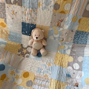 Könnte beinhalten: Ein Patchwork-Quilt in Gelb-, Blau-, Grau- und Weißtönen mit Mond-, Stern- und Bärenmustern. Ein beiger Teddybär mit braunen Details liegt auf dem Quilt. Der Quilt ist für ein Baby konzipiert, mit Maßen in Zentimetern.