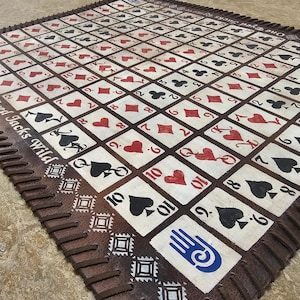 May include: A handmade playing card game mat featuring a grid of playing card suits in red, black, and white. The mat has a brown leather border with the text "Two Eyed Jacks Wild" and decorative stitching. The mat is designed for card games.