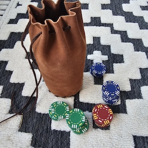 Puede incluir: Una bolsa de gamuza marrón con cordón y fichas de póquer. Las fichas son verdes, azules y rojas con detalles blancos. La bolsa está sobre una alfombra con estampado en blanco y negro.