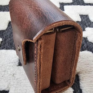 May include: A brown leather case with a textured surface and a flap closure. The case has a rectangular shape with rounded edges and a lighter brown interior. The leather has a distressed appearance.