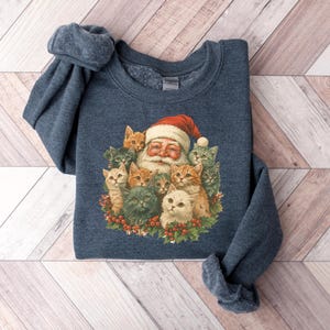 May include: A navy blue sweatshirt featuring a festive Santa Claus surrounded by a group of adorable cats. Santa wears a red hat and is encircled by various colored cats. The design is printed on a soft, comfortable sweatshirt.