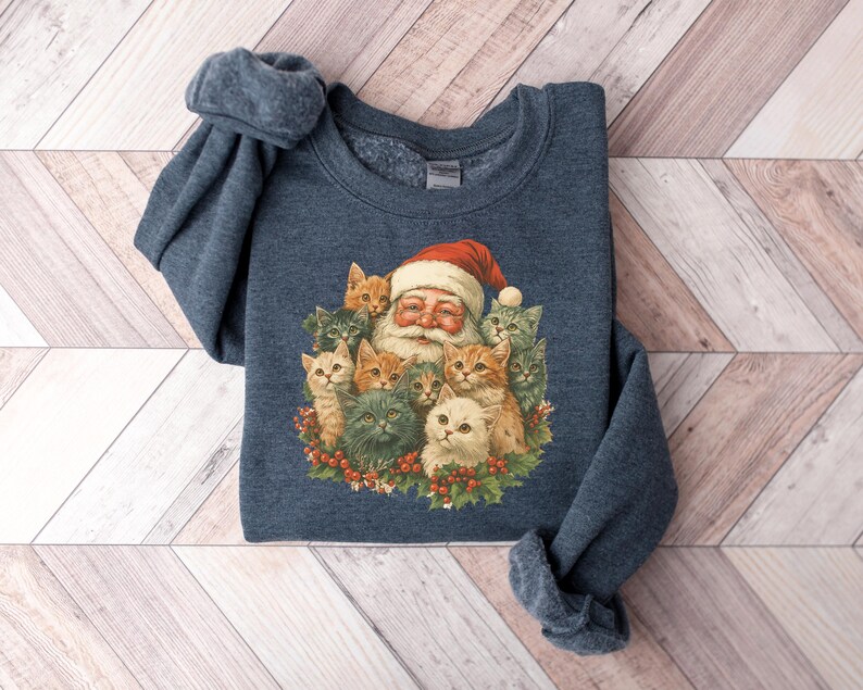 May include: A navy blue sweatshirt featuring a festive Santa Claus surrounded by a group of adorable cats. Santa wears a red hat and is encircled by various colored cats. The design is printed on a soft, comfortable sweatshirt.