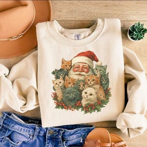 May include: Cream-colored sweatshirt featuring a graphic of Santa Claus surrounded by a group of cats. The Santa illustration has a red hat and white beard. The cats are various colors, including orange, gray, and white.