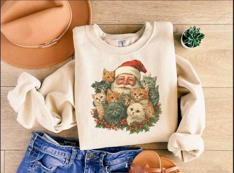 May include: Cream-colored sweatshirt featuring a graphic of Santa Claus surrounded by a group of cats. The Santa illustration has a red hat and white beard. The cats are various colors, including orange, gray, and white.