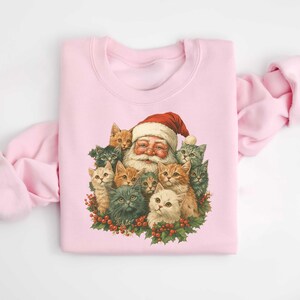 May include: A light pink sweatshirt featuring a vintage illustration of Santa Claus surrounded by several cats. Santa is wearing a red hat and smiling. The cats are various colors, including gray, orange, and white. The cats and Santa are surrounded by a wreath of holly.