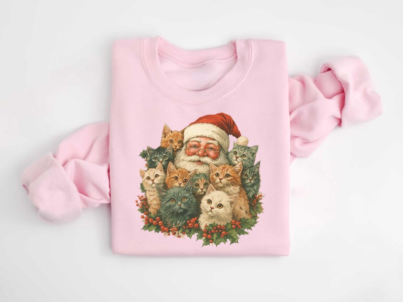 May include: A light pink sweatshirt featuring a vintage illustration of Santa Claus surrounded by several cats. Santa is wearing a red hat and smiling. The cats are various colors, including gray, orange, and white. The cats and Santa are surrounded by a wreath of holly.