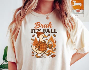 May include: Cream-colored t-shirt with the text "Bruh It's Fall" in brown and black. The graphic features a cartoon duck wearing a hat and plaid shirt, holding a coffee cup, surrounded by fall leaves.