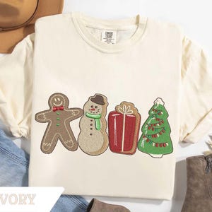 May include: An ivory short-sleeve t-shirt featuring an embroidered design of a gingerbread person with a red bow tie, a snowman wearing a brown hat and green scarf, a red gift box with a white bow, and a green Christmas tree with colorful lights.