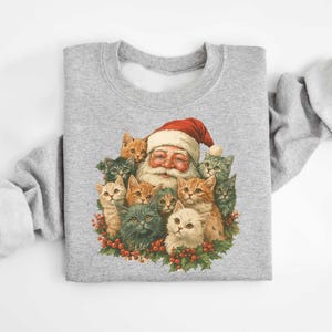 May include: A light gray sweatshirt featuring a festive graphic of Santa Claus surrounded by a group of adorable kittens. Santa is wearing his traditional red hat and has a long white beard. The kittens are in various colors, including orange, gray, and white.