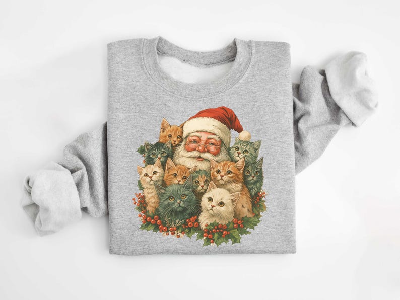 May include: A light gray sweatshirt featuring a festive graphic of Santa Claus surrounded by a group of adorable kittens. Santa is wearing his traditional red hat and has a long white beard. The kittens are in various colors, including orange, gray, and white.