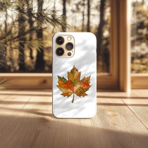 May include: A white phone case with a colourful maple leaf design. The leaf is made up of various autumn colours, including orange, green, and red, creating a patchwork effect. The case is on a wooden surface.