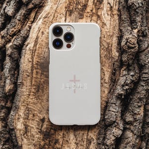 May include: A light gray phone case with a pink cross and the words "Jesus my life" printed on the back. The phone case has a camera cutout and is set against a textured, brown wooden background.