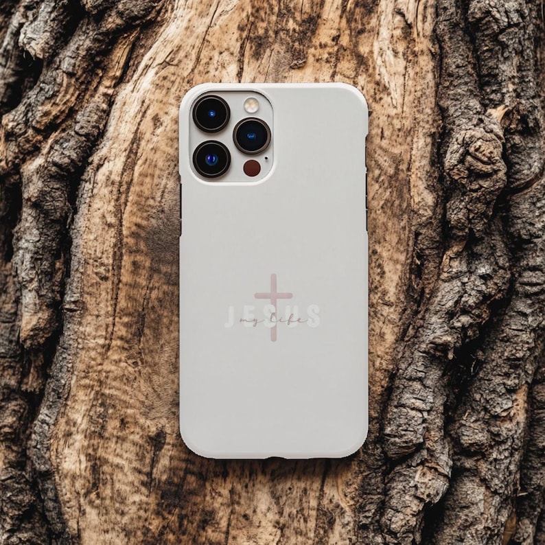 May include: A light gray phone case with a pink cross and the words "Jesus my life" printed on the back. The phone case has a camera cutout and is set against a textured, brown wooden background.