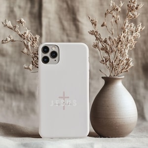 May include: A white phone case with the words "Jesus my life" and a cross graphic in a soft pink color. The phone case is next to a vase with dried flowers.